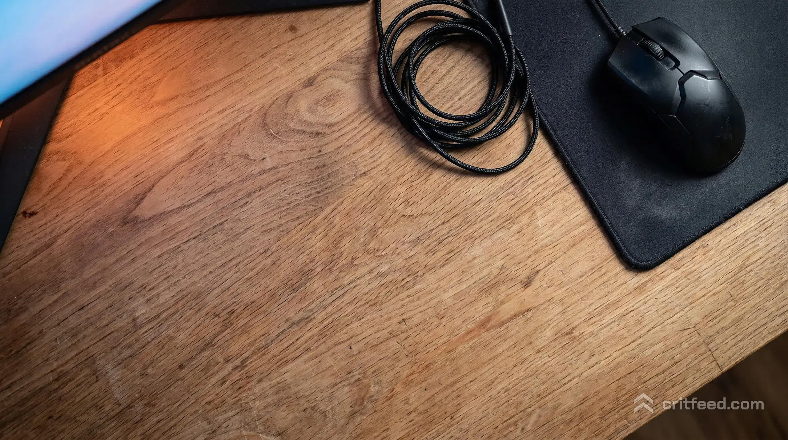 an overhead shot of a wooden desk with a gaming mouse pushed to one side, a coiled headset cable, and a monitor glowing just