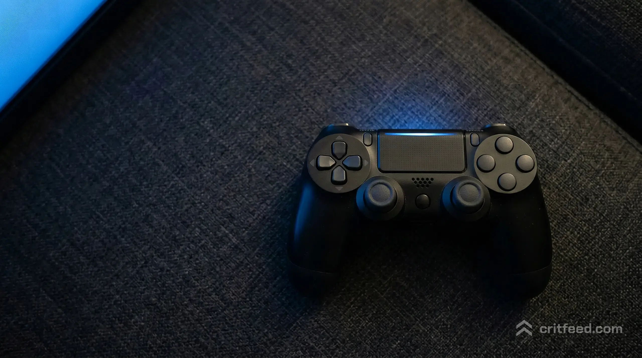 a PS4 controller resting on a dark fabric couch, the home screen glow from a television washing the plastic in soft blue