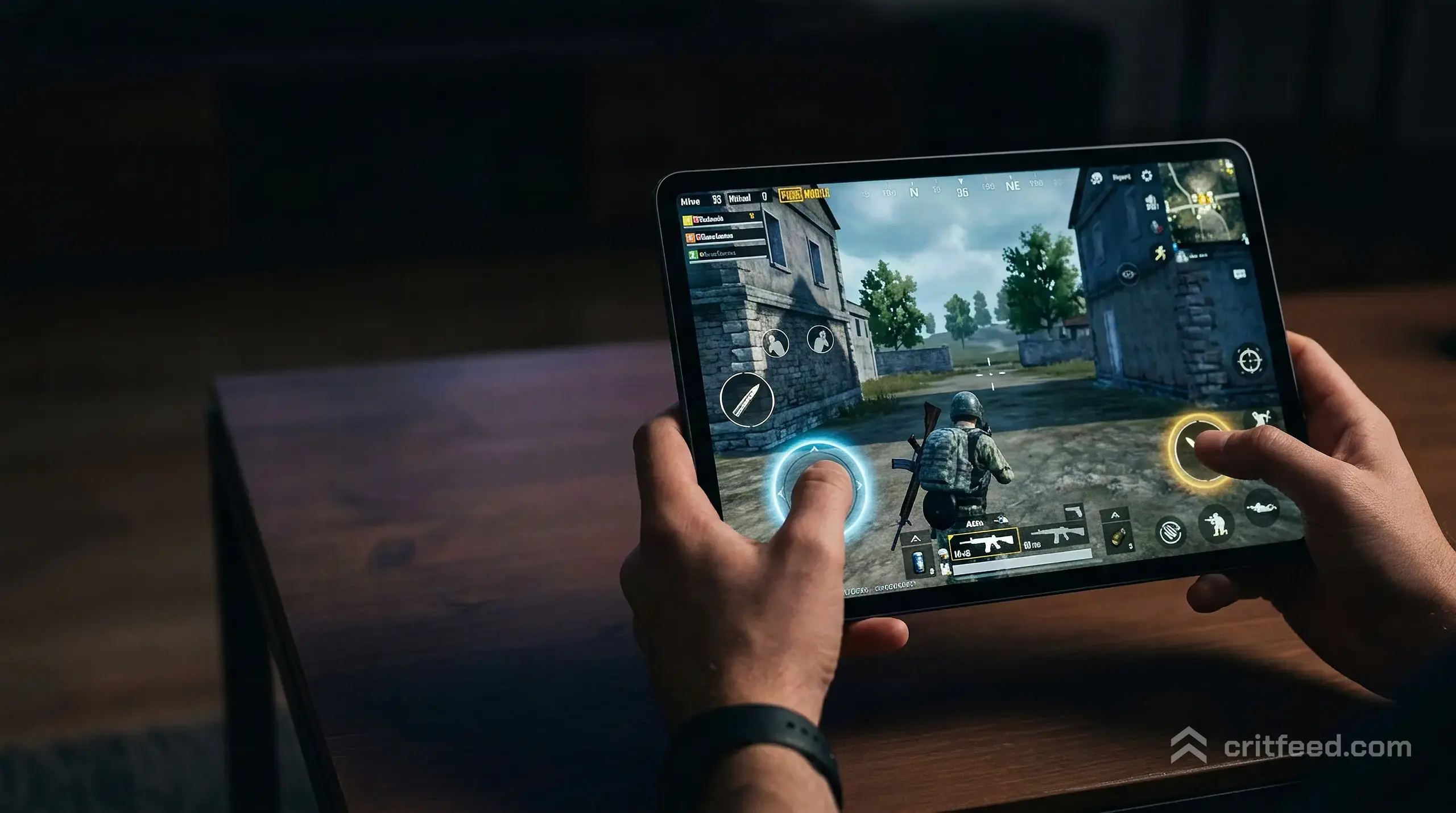 A pair of hands on a large tablet in landscape, thumbs resting on the on-screen fire and movement buttons, the room lit only
